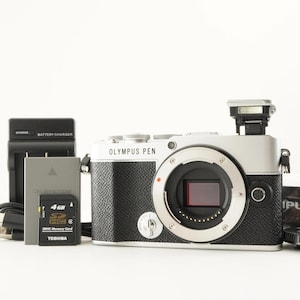 Può includere: Una fotocamera digitale Olympus Pen argento e nera con flash montato, accompagnata da un caricabatterie, una batteria grigia e una scheda di memoria SDHC Toshiba da 4 GB. Il corpo presenta un'impugnatura nera testurizzata e un innesto obiettivo.