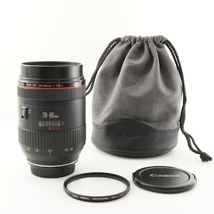 Puede incluir: Un objetivo de cámara Canon EF 28-80mm con una tapa de objetivo, un filtro protector y una bolsa gris con cordón. El objetivo es negro con marcas rojas y blancas. La tapa y la bolsa tienen el logotipo de Canon.