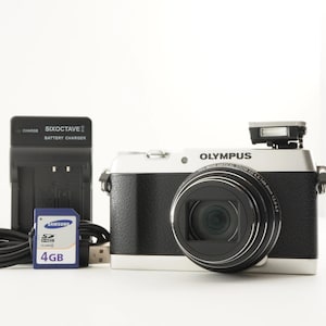 Può includere: Una fotocamera digitale Olympus argento e nera con obiettivo, flash a scomparsa, un caricabatterie nero Sixoctave con cavo e una scheda di memoria SDHC Samsung da 4GB blu. L'obiettivo riporta la scritta 24x wide optical zoom ED 4.5-108.0mm.