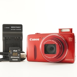 May include: A red Canon SX600 HS digital camera with a 18x optical zoom lens, a battery charger, and a 4GB SDHC memory card. The camera has the Canon logo on the front.