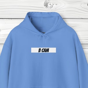 Może przedstawiać: Jasna niebieska bluza z kapturem z białym prostokątem z przodu, na którym widnieje napis "B CAM" czarnym tekstem.