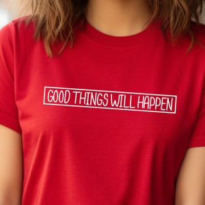 May include: A red t-shirt with the phrase "GOOD THINGS WILL HAPPEN" in white, inside a white rectangle. The shirt has short sleeves and a crew neck.