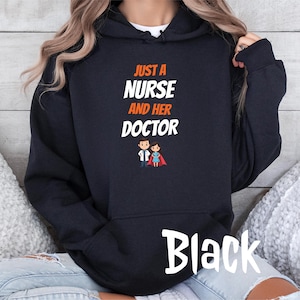 Könnte beinhalten: Schwarzer Kapuzenpullover mit dem Text "JUST A NURSE AND HER DOCTOR" in Weiß und Orange. Das Design beinhaltet eine Cartoon-Krankenschwester und einen Arzt. Das Wort "Black" ist in einer Schreibschrift am unteren Rand des Hoodies geschrieben.