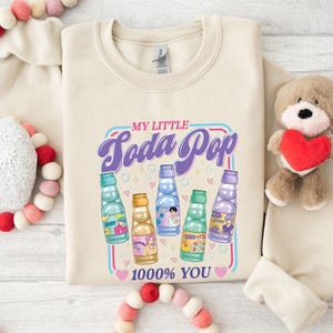 May include: Cream-colored sweatshirt with a graphic that reads "MY LITTLE Soda Pop" in purple and blue. The graphic features cartoon soda bottles in various colors. The text "1000% YOU" is below the bottles.