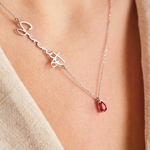 May include: A silver necklace with a cursive name pendant and a teardrop-shaped red gemstone pendant.