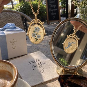 May include: A gold-toned oval pendant necklace featuring a rotating center piece. One side displays a blue forget-me-not flower illustration on a textured gold background, while the reflection in a round mirror shows the letter F in blue script.
