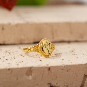 Puede incluir: Un anillo dorado con una cara ovalada con un diseño floral negro. El anillo tiene una banda detallada con un diseño ornamentado y un acabado pulido. El anillo se muestra sobre una superficie clara y texturizada.