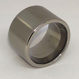 Op de afbeelding: Een brede, cilindrische ring gemaakt van geborsteld titanium. De ring heeft een gladde, matte afwerking en een gepolijst interieur. Het woord "TITANIUM" is in de band gegraveerd. De ring is zilvergrijs van kleur.