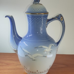 Puede incluir: Una cafetera de porcelana azul y blanca con tapa y asa. La cafetera presenta un diseño de gaviotas volando y ribetes dorados. La tapa está rematada con un adorno decorativo. La cafetera es probablemente vintage.