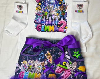 Może przedstawiać: Biały t-shirt z grafiką urodzinową Chuck E. Cheese, napisem BIRTHDAY GIRL, GEMMA oraz cyfrą 2. Zestaw zawiera białe skarpetki z imieniem GEMMA oraz ciemną dżinsową spódniczkę z fioletową wstążką, fioletowymi piórami i nadrukami postaci.