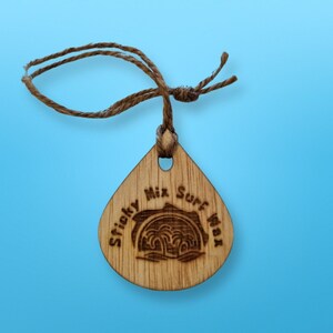 May include: A wooden surfboard wax tag with a brown twine string. The tag is shaped like a teardrop and has the text "Sticky Mix Surf Wax" engraved on it.