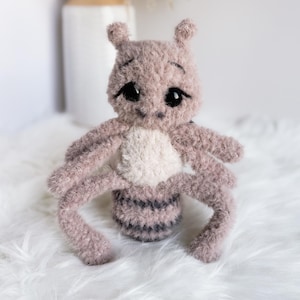 May include: A handmade plush toy insect in shades of mauve, cream, and gray. The toy has large, black eyes, a cream-colored chest, and a striped abdomen. The insect has six legs and two antennae.