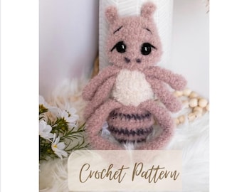 May include: A crocheted amigurumi toy in the shape of a pink and white bug with large black eyes. The toy is sitting on a white fluffy surface. The text "Crochet Pattern" is visible at the bottom of the image.