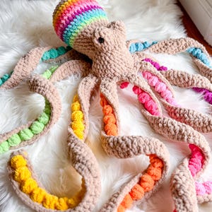 May include: A large, handmade crochet octopus in shades of tan, with a rainbow-colored head. The tentacles are tan with colorful pom-pom accents in green, yellow, orange, pink, and blue. The plush toy is displayed on a white, fluffy surface.