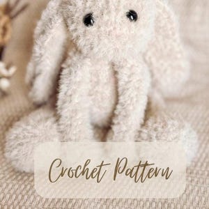 May include: A crocheted bunny rabbit toy with a fluffy white texture and black eyes. The bunny is sitting on a beige fabric surface. The text "Crochet Pattern" is visible below the bunny.