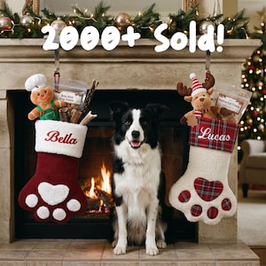 May include: Two paw-shaped Christmas stockings hanging from a fireplace mantel. The left stocking is maroon with the name Bella and a gingerbread plush. The right stocking is white with a plaid cuff, the name Lucas, and a reindeer plush.