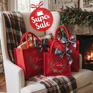 May include: Three red tote bags with sturdy handles, each featuring a printed wreath design and a personalized name in white script. The bags are adorned with festive patterned bows and are displayed together on a white upholstered chair.