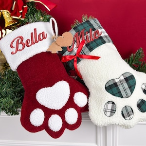 May include: Two Christmas stockings shaped like pet paws. One is burgundy with a white heart and paw pads, the other is white with a green plaid paw print. The names "Bella" and "Milo" are embroidered on the cuffs.