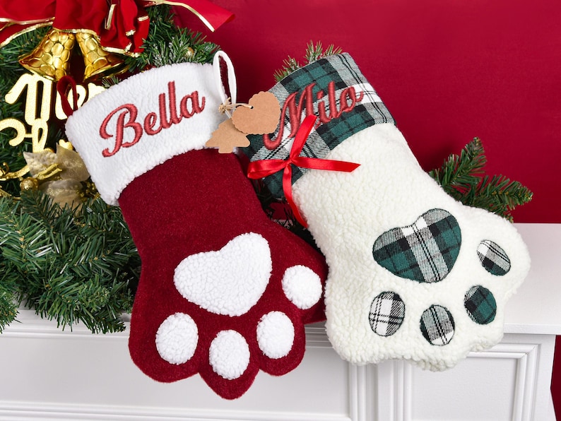 May include: Two paw-shaped Christmas stockings. The left stocking is deep red with a white cuff embroidered with the name Bella. The right stocking is white with a green and black plaid cuff embroidered with the name Milo and a red ribbon bow.