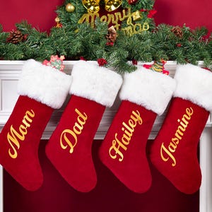 May include: Four red Christmas stockings with white faux fur cuffs hanging in a row. Each stocking features gold cursive lettering reading Mom, Dad, Hailey, and Jasmine. The stockings are displayed against a white fireplace mantel.