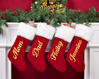 May include: Four red Christmas stockings with white faux fur cuffs hanging in a row. Each stocking features gold cursive lettering reading Mom, Dad, Hailey, and Jasmine. The stockings are displayed against a white fireplace mantel.