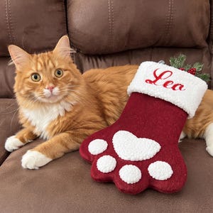 May include: A maroon felt pet stocking shaped like a paw print with a white sherpa cuff embroidered with the name Leo in red script. The stocking features a large white sherpa heart-shaped pad and four smaller white sherpa toe pads.