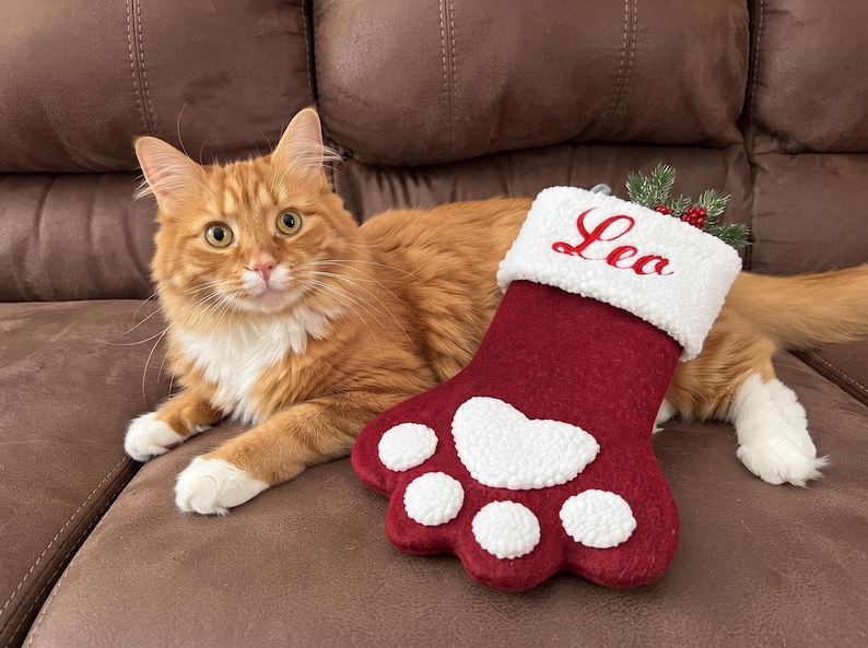 May include: A maroon felt pet stocking shaped like a paw print with a white sherpa cuff embroidered with the name Leo in red script. The stocking features a large white sherpa heart-shaped pad and four smaller white sherpa toe pads.