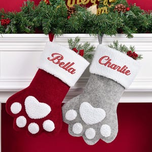 May include: Two paw-shaped Christmas stockings, one deep red and one light gray, each featuring a white sherpa cuff embroidered with the names Bella and Charlie in red thread. The stockings have white sherpa paw pads and toe accents.