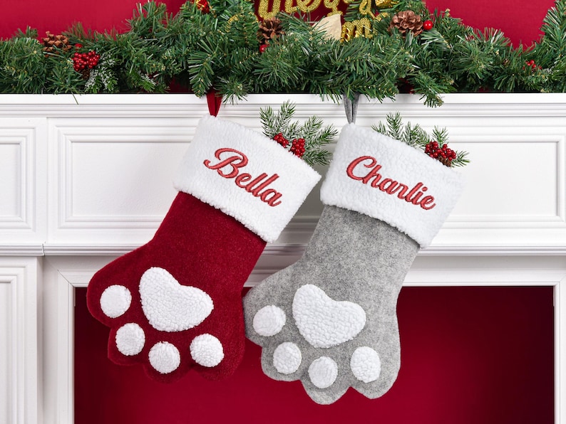 May include: Two paw-shaped Christmas stockings, one deep red and one light gray, each featuring a white sherpa cuff embroidered with the names Bella and Charlie in red thread. The stockings have white sherpa paw pads and toe accents.