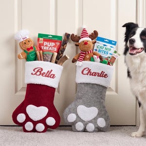 May include: Two paw-shaped Christmas stockings, one deep red and one grey, each featuring a white cuff embroidered with the names Bella and Charlie. The stockings are filled with various pet toys and treat bags, including a gingerbread man and a reindeer.