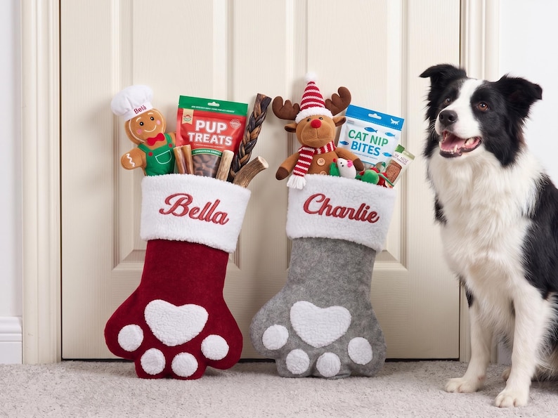 May include: Two paw-shaped Christmas stockings, one deep red and one grey, each featuring a white cuff embroidered with the names Bella and Charlie. The stockings are filled with various pet toys and treat bags, including a gingerbread man and a reindeer.