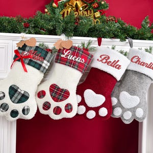 May include: Four paw-shaped Christmas stockings with personalized names embroidered on the cuffs. The first is white with a green plaid cuff and heart pad, the second is white with a red plaid cuff and heart pad, the third is red with a white cuff, and the fourth is gray with a white cuff.