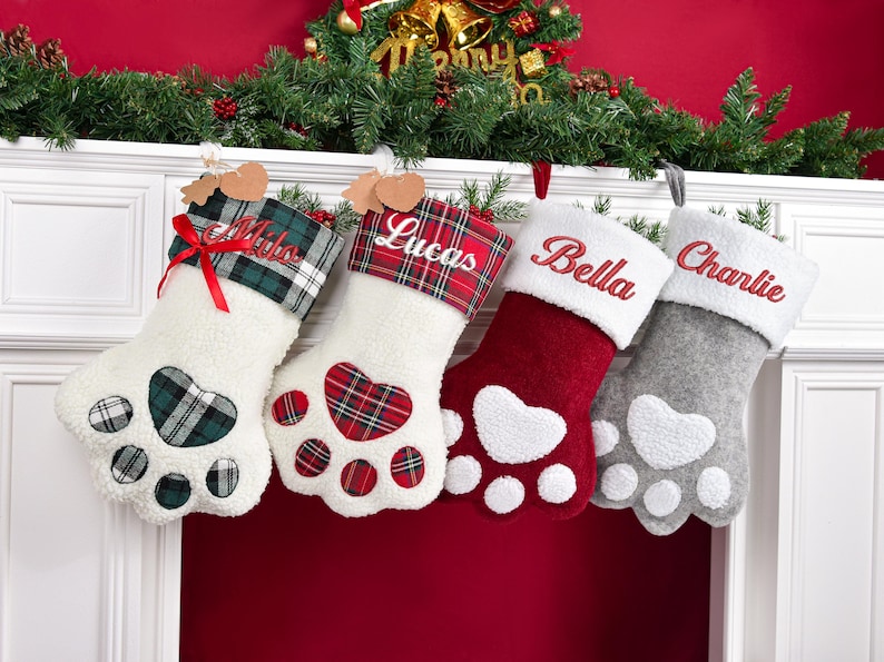 May include: Four paw-shaped Christmas stockings with personalized names embroidered on the cuffs. The first is white with a green plaid cuff and heart pad, the second is white with a red plaid cuff and heart pad, the third is red with a white cuff, and the fourth is gray with a white cuff.