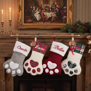 May include: Four paw-shaped Christmas stockings hanging on a rod. The first is gray with a white cuff and paw pad. The second is white with a red plaid cuff and paw pad. The third is red with a white cuff and paw pad. The fourth is white with a green plaid cuff and paw pad. Each features embroidered names: Charlie, Lucas, Bella, and Olivia.