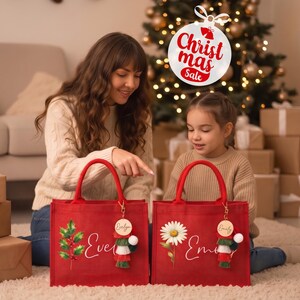 May include: Two red tote bags with sturdy handles, each featuring a personalized name in white script, a decorative wooden tag, and a colorful tassel. One bag displays a holly branch graphic, while the other features a white daisy illustration.
