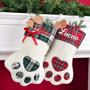 May include: Two white sherpa paw-shaped Christmas stockings. The left stocking features a green plaid cuff with the name Milo embroidered in red and a red ribbon bow. The right stocking has a red plaid cuff with the name Lucas embroidered in white.