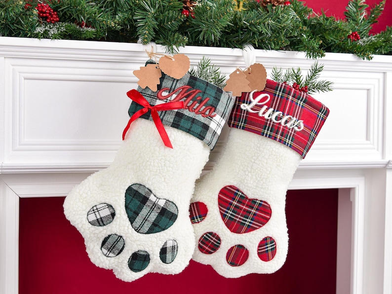 May include: Two white sherpa paw-shaped Christmas stockings. The left stocking features a green plaid cuff with the name Milo embroidered in red and a red ribbon bow. The right stocking has a red plaid cuff with the name Lucas embroidered in white.