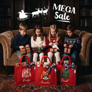 May include: Three red tote bags with handles, each featuring a printed holiday sweater graphic and a personalized name in white script. The bags are labeled Evelyn, Amelia, and Jacob, and each is decorated with a patterned ribbon tied to the handle.