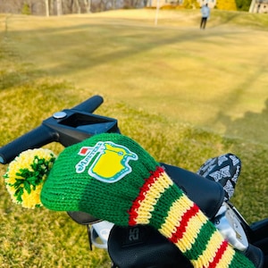 May include: Green, yellow, and red knitted golf club head cover with a Masters logo patch and a yellow and green pom-pom.