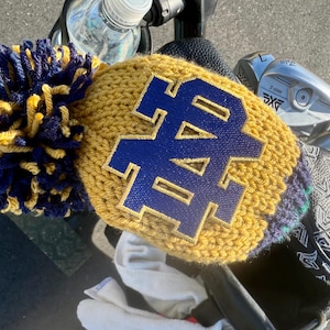 May include: A gold and navy blue knitted golf club head cover with a large pom-pom. The cover features the letters "ND" in navy blue. Other golf club covers and golf clubs are visible in the background.