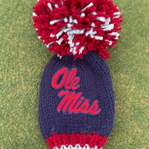 May include: A navy blue knit golf club headcover with the words "Ole Miss" in red script. The headcover has a red and white pom-pom and a red and white striped cuff. The headcover is on a green surface.