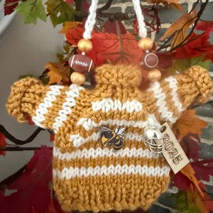 May include: Hand-knitted, mustard yellow and white striped sweater ornament with a bee pin and a miniature football helmet charm. The ornament is suspended by a white cord with wooden beads and football-shaped accents. A wooden tag with "2025" hangs from the ornament.