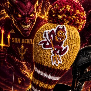 May include: A golf club head cover with a yellow, maroon, and white striped design, featuring a cartoon devil logo. The background includes a menacing devil face and the words "Sun Devils". A pom-pom tops the cover.
