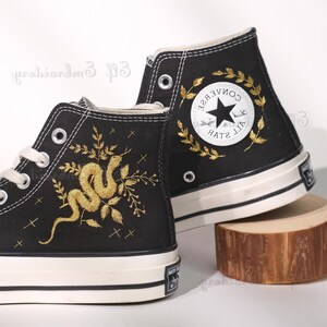 May include: Black high-top Converse sneakers with gold embroidery of a snake and leaves on one shoe and a gold embroidered wreath around the Converse All Star logo on the other shoe.