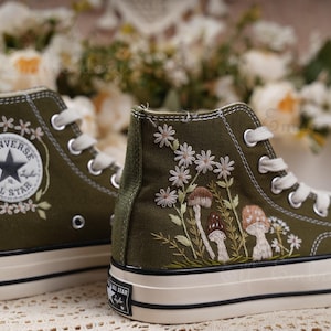 May include: A pair of green canvas high-top sneakers with white soles and laces. The sneakers are embroidered with flowers and mushrooms. The left sneaker has the Converse All Star logo.