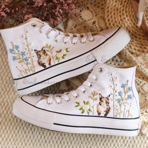 Puede incluir: Zapatillas blancas de lona de caña alta con diseños florales bordados y un gato. El gato es marrón, blanco y naranja con ojos azules.