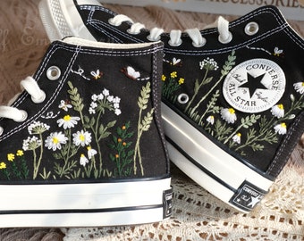 Könnte beinhalten: Ein Paar schwarze High-Top-Canvas-Sneaker mit weißen Sohlen. Die Sneaker sind mit weißen Blumen, grünen Blättern und gelben Akzenten bestickt. Der linke Sneaker hat das Converse-Logo und den Text "Chuck Taylor All Star" an der Seite.