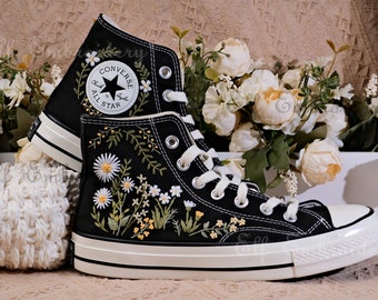 Puede incluir: Zapatillas Converse negras de caña alta con costuras blancas y suela de goma blanca. Las zapatillas están bordadas con margaritas blancas, hojas verdes y flores amarillas. El logotipo de Converse es visible en el lateral de la zapatilla.