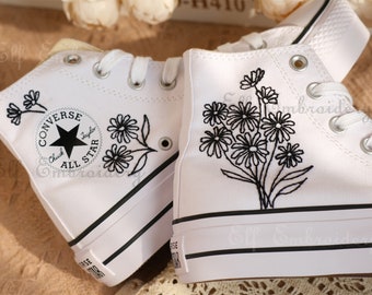 May include: White high-top Converse sneakers with black embroidered flowers and the Converse All Star logo on the side.