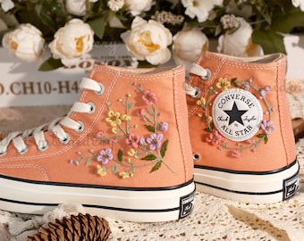 May include: A pair of high-top canvas sneakers with embroidered floral designs. The sneakers are peach-colored with white soles and black accents. The Converse All Star logo is visible on the side of one shoe.